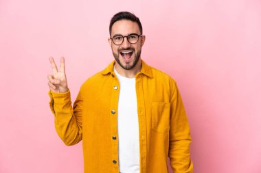 Beyaz tenli yakışıklı adam pembe arka planda izole edilmiş gülümsüyor ve zafer işareti gösteriyor.