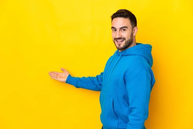 Sarı arka planda izole edilmiş beyaz tenli yakışıklı adam, davet edilmek için ellerini yana uzatıyor.