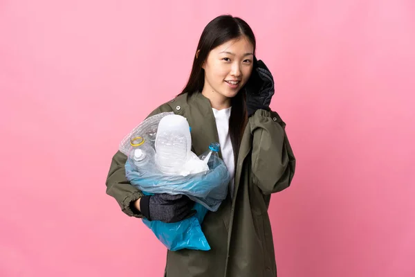 Chinese girl holding a bag full of plastic bottles to recycle over isolated pink background keeping a conversation with the mobile phone with someone