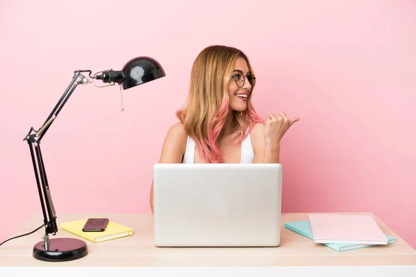 Young student woman in a workplace with a laptop over pink background pointing to the side to present a product