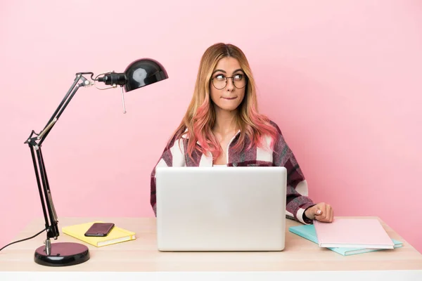 Young student woman in a workplace with a laptop over pink background having doubts while looking up