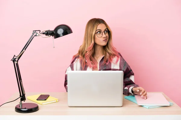 Young student woman in a workplace with a laptop over pink background making doubts gesture looking side