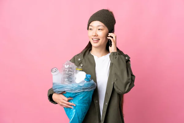 Chinese girl holding a bag full of plastic bottles to recycle over isolated pink background keeping a conversation with the mobile phone