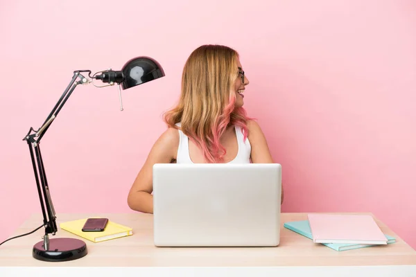 Young student woman in a workplace with a laptop over pink background laughing in lateral position