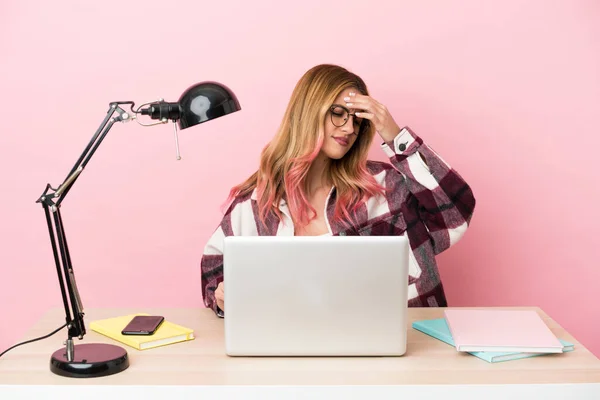 Young student woman in a workplace with a laptop over pink background with headache