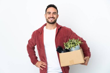 Beyaz arka planda izole edilmiş eşyalarla dolu bir kutu alıp kalçasında kollarıyla poz veren ve gülümseyen genç beyaz bir adam hamle yapıyor.