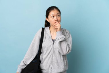 İzole edilmiş mavi arka planda spor çantalı genç Çinli kadının şüpheleri ve kafa karıştırıcı bir yüz ifadesi var.