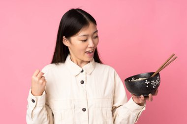 Pembe arka planda izole edilmiş genç Asyalı bir kadın zaferi kutlarken elinde yemek çubuklarıyla bir kase erişte tutuyor.