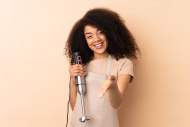 Genç Afrikalı Amerikalı kadın el blenderi kullanıyor. İyi bir anlaşmadan sonra bej arka planda el sıkışırken izole edilmiş.