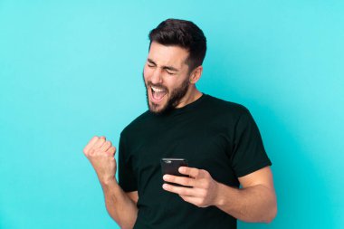 Beyaz tenli yakışıklı adam mavi arka planda izole edilmiş ve telefon zafer pozisyonunda.