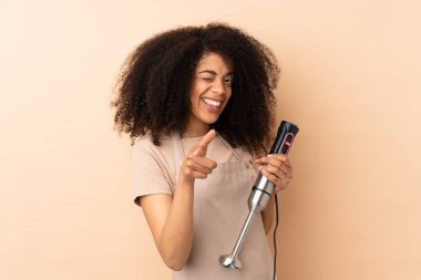 Genç Afrikalı Amerikalı kadın bej arka planda izole edilmiş el blenderi kullanıyor.