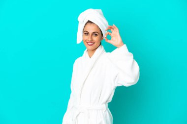 İzole edilmiş mavi arka planda bornozlu genç bir kadın parmaklarıyla tamam işareti gösteriyor.