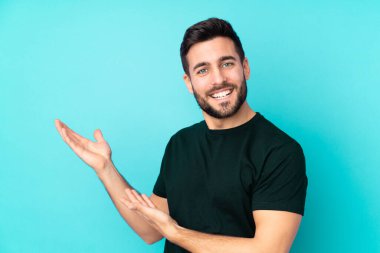 Beyaz tenli yakışıklı adam mavi arka planda izole edilmiş. Davet etmek için ellerini yana uzatıyor.