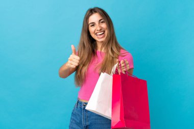 Soyutlanmış mavi arka planda genç bir kadın elinde alışveriş torbalarıyla ve başparmağıyla