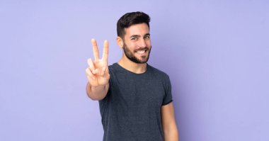 Beyaz tenli yakışıklı adam gülümsüyor ve izole edilmiş mor arka planda zafer işareti gösteriyor.