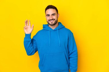 Beyaz tenli yakışıklı adam sarı arka planda izole edilmiş mutlu bir ifadeyle selamlıyor.