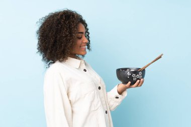Genç Afrikalı Amerikalı bir kadın, elinde çubuklarla elinde bir kase erişte tutarken mutlu bir ifadeyle mavi arka planda izole edilmiş.