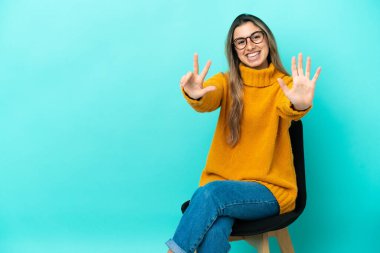 Mavi arka planda izole edilmiş bir sandalyede oturan beyaz bir kadın sekiz parmaklı sayıyor.