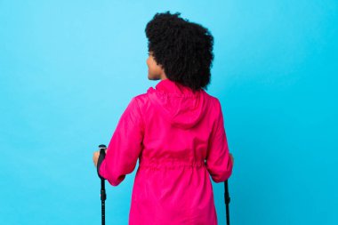 Sırt çantalı ve yürüyüş direkleriyle genç Afrikalı Amerikalı mavi arka planda izole edilmiş ve yan tarafa bakıyor.