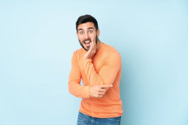 Beyaz, yakışıklı bir adam bir ürün sunmak için kenarı işaret ediyor ve izole edilmiş mavi arka plan üzerine bir şeyler fısıldıyor.