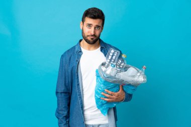 Mavi arka planda izole edilmiş bir şekilde geri dönüşüm için plastik şişe dolu bir çanta taşıyan genç beyaz bir adam.