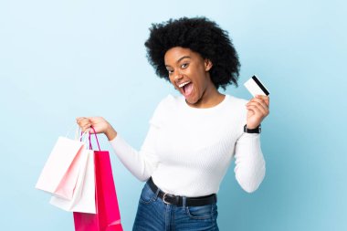 Genç Afrikalı Amerikalı bir kadın mavi arka planda izole edilmiş. Elinde alışveriş çantaları ve bir kredi kartı var.