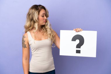 Mor arka planda izole edilmiş genç Brezilyalı bir kadın soru işareti sembollü bir levha tutuyor.