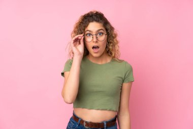 Pembe arka planda gözlüklü, kıvırcık saçlı ve şaşırmış genç sarışın kadın.