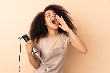 El blenderi kullanan Afrikalı genç bir kadın bej arka planda ağzı açık bağırıyor.