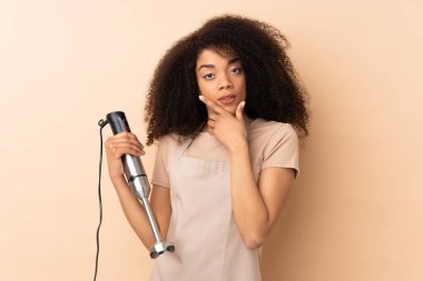 Genç Afrikalı Amerikalı kadın, bej arka planda izole edilmiş bir el blenderi kullanıyor.