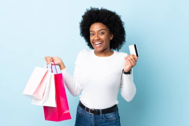 Genç Afrikalı Amerikalı bir kadın mavi arka planda izole edilmiş. Elinde alışveriş çantaları ve bir kredi kartı var.