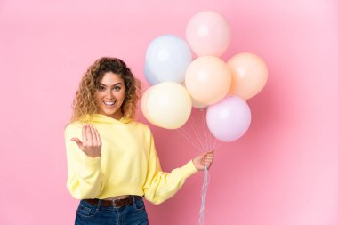 Kıvırcık saçlı genç bir kadın pembe arka planda izole edilmiş bir sürü balon yakalıyor.