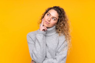 Sarışın, kıvırcık saçlı, balıkçı yaka kazak giyen genç bir kadın sarı arka planda izole edilmiş bir şekilde yukarı bakarken bir fikir düşünüyor.