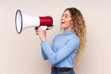 Sarışın, kıvırcık saçlı, balıkçı yaka kazak giyen, bej arka planda izole edilmiş, megafonla bağıran genç bir kadın.