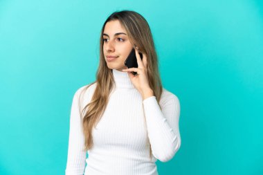 Mavi arka planda izole edilmiş cep telefonu kullanan genç beyaz kadın yan tarafa bakıyor.