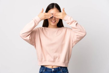 İzole edilmiş beyaz arka planı olan genç bir Çinli kız elleriyle gözlerini kapatıyor ve gülümsüyor.