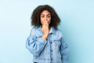 Genç Afrikalı Amerikalı bir kadın, mavi arka planda izole edilmiş, ağzı elle kapatılmış.