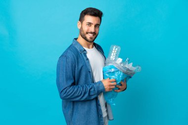 Mavi arka planda izole edilmiş plastik şişelerle dolu bir çanta taşıyan genç beyaz bir adam çok fazla gülümsüyor.