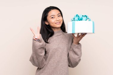 Genç Asyalı kız elinde büyük bir pasta tutuyor. Bej arka planda izole edilmiş. Gülümsüyor ve zafer işareti gösteriyor.