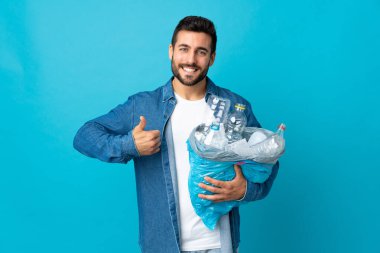 Elinde plastik şişelerle dolu bir çanta tutan genç beyaz bir adam mavi arka planda izole edilmiş bir şekilde geri dönüşüm yapıyor.