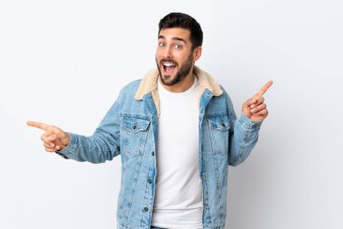 Beyaz arka planda sakallı genç yakışıklı adam parmak uçlarını işaret ediyor ve mutlu.