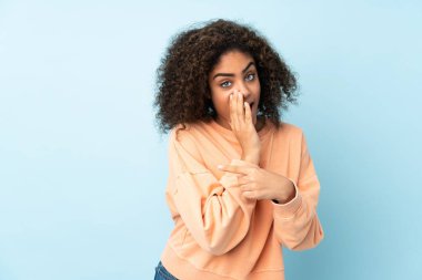 Mavi arka planda izole edilmiş genç Afrikalı Amerikalı kadın bir ürünü sunmak ve bir şeyler fısıldamak için kenarı işaret ediyor.