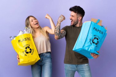 Mor arka planda izole edilmiş, kazanan pozisyonda zaferi kutlayan genç beyaz çift geri dönüşüm yapıyor.