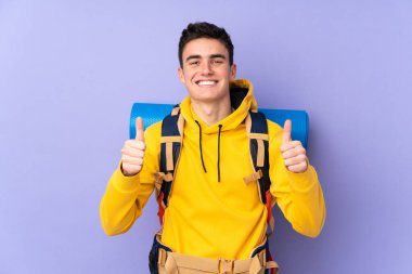 Mor arka planda izole edilmiş büyük sırt çantalı Kafkasyalı genç bir dağcı baş parmak işareti yapıyor.