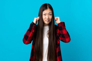 Genç Çinli kız, izole edilmiş mavi arka planda hayal kırıklığına uğramış ve kulaklarını kapatmış.