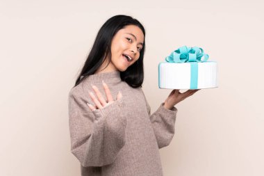 Genç Asyalı kız elinde büyük bir pasta tutarak bej arka planda izole edilmiş mutlu bir ifadeyle selamlıyor.