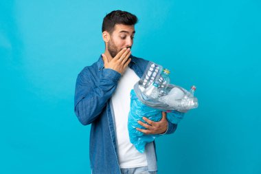 Elinde plastik şişelerle dolu bir çanta tutan beyaz bir adam mavi arka planda izole edilmiş, şaşırmış ve şok olmuş bir yüz ifadesiyle.