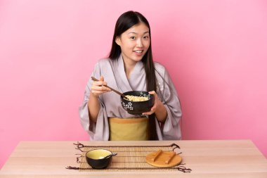 Genç Çinli kız kimono giyiyor ve pembe arka planda bir masada erişte yiyor.