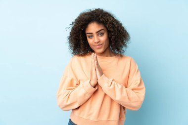 Mavi arka planda izole edilmiş genç Afrikalı Amerikalı kadın avuç içlerini bir arada tutuyor. İnsan bir şey ister.
