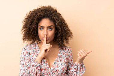 Genç Afro-Amerikalı kadın bej arka planda izole edilmiş. Yan tarafı işaret ediyor ve sessizlik hareketi yapıyor.